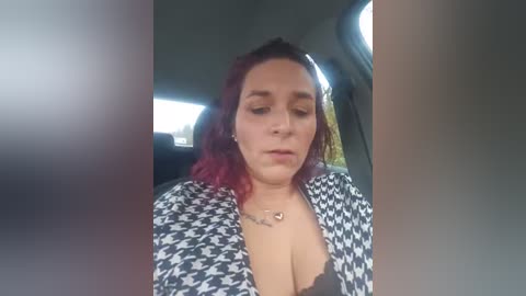 Video of a plus-size, light-skinned woman with shoulder-length red hair, wearing a black-and-white houndstooth jacket and black lace bra, sitting in a car with dark tinted windows.