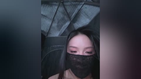 A video of an East Asian woman with long black hair and light skin, wearing a black lace face mask, standing in a dimly lit, industrial room with metallic ceiling panels.