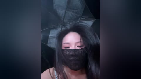 Video of an Asian woman with long black hair, wearing a black mask and holding a black umbrella. The background is dark and indistinct, suggesting an indoor setting.