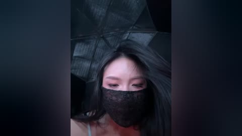 Video of a woman with long black hair, wearing a black lace mask, standing under an open black umbrella in a dark, shadowy environment.