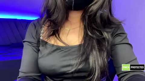 A video of a woman with long, wavy black hair, wearing a black face mask, black long-sleeve top, and a gold necklace. Background features a purple and blue lit room, with a watermark \"ProTecter\" at the bottom right.