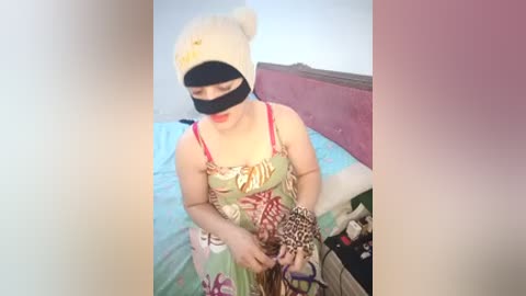 Video of a woman in a leopard print dress, holding a leopard print purse, wearing a black mask, and a white teddy bear hat, in a bedroom with a pink headboard and blue bedding.