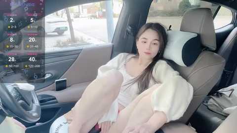 A video of a young woman with long hair, wearing a white sweater, sitting in a car with the door open, outside in daylight.