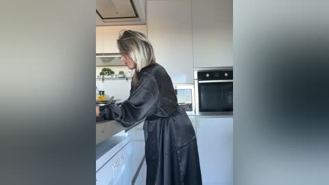 Video of a woman with blonde hair in a black robe standing in a modern, minimalist kitchen with white cabinets, stainless steel appliances, and a sleek countertop.