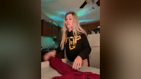 A candid video of a blonde woman in a black sweatshirt with yellow text, seated at a kitchen counter, pulling a red blanket from a pile.