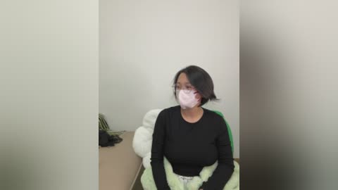 Video of an Asian woman with short black hair, wearing a black long-sleeve shirt, a white surgical mask, and a light green blanket over her lap. She sits on a beige couch in a plain, white-walled room.