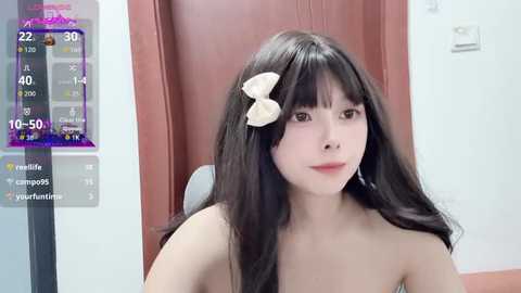 A video of a young Asian woman with long black hair, wearing a white bow, and a bare chest, taken indoors with a temperature display in the background.