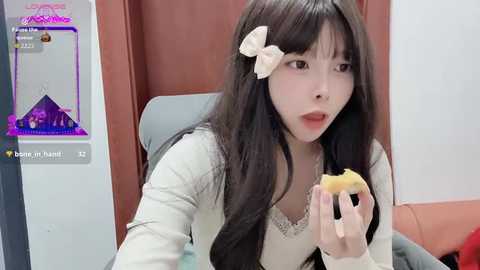 Video of a young Asian woman with long black hair, wearing a white bow, eating a donut in a cozy, dimly lit room with a pink and purple aesthetic.