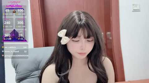 A video of a young Asian woman with long, dark hair, wearing a white bow, sitting indoors, looking down.
