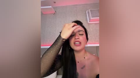 Video of a young woman with long black hair, wearing a black, sparkly long-sleeve top, standing indoors with a hand on her forehead, mouth open, against a textured beige wall with red lighting.