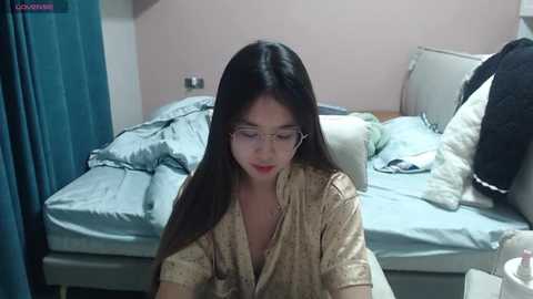 Video of an Asian woman with long black hair, wearing glasses and a beige robe, sitting on a bed with blue sheets, surrounded by pillows.