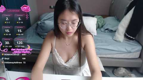 A video of an Asian woman with long black hair and glasses, wearing a white lace camisole, sitting at a table in a messy bedroom.