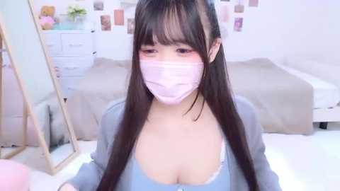 A video of an Asian woman with long black hair, wearing a pink face mask, blue top, and large breasts, in a pastel-colored bedroom with a bed and white furniture.