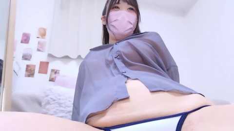 Video of an Asian woman with fair skin, wearing a gray shirt, white panties, and a pink face mask, lying on a bed in a minimalist room.