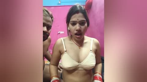 Video of a South Asian woman with medium skin tone, wearing a beige bra, red bangles, and bindi. She has a distressed expression, surrounded by two men in the background. The setting is a vibrant pink room.