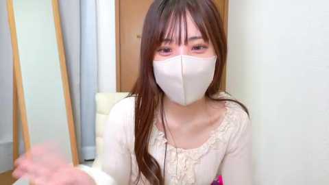 Video of an East Asian woman with long brown hair and light skin, wearing a white mask, white blouse, and a pink top, indoors, looking directly at the camera.