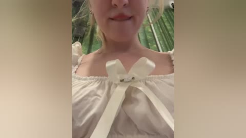 Video of a smiling woman with light skin, wearing a white off-the-shoulder dress with a large bow at the neckline, standing in front of green foliage.