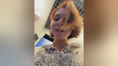 Video of a young person with short, messy red hair, wearing a patterned hoodie, smiling slightly in a brightly lit room with a blue and black tiled wall and a framed picture in the background.