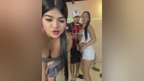 Video of three young women in a dimly lit room. One woman, with long black hair, leans forward, wearing a revealing gray top, while the other two, wearing casual outfits, stand behind her.