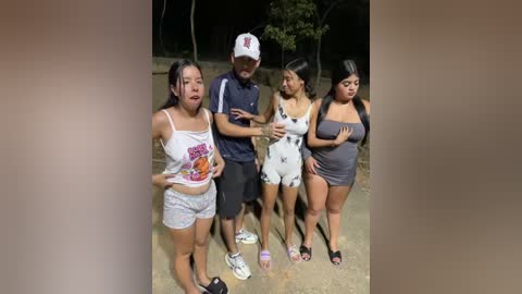 Video of four women and a man outdoors at night, all wearing casual attire, standing closely together, with a man in a white cap and a woman in a Hooters shirt.