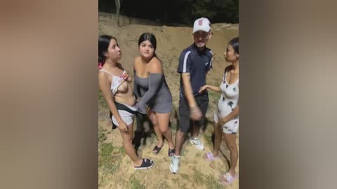 A video shows four people, including a woman in a gray dress and another in a white bikini, standing on a sandy ground. A man in a baseball cap and a woman in a white dress are also present.