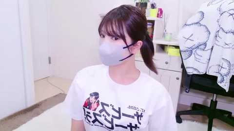 Video of an East Asian woman with light skin, dark hair in a ponytail, wearing a white T-shirt with a graphic, gray face mask, sitting in a white room with a black office chair and a white door.