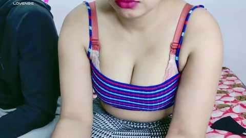 Video of a woman with fair skin, wearing a blue and pink striped crop top, exposing ample cleavage, sitting on a patterned bedspread in a bedroom.