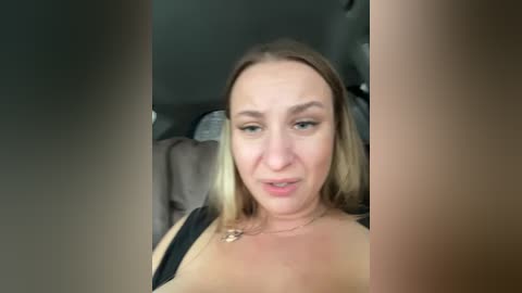 Video of a blonde woman with light skin, wearing a black top, seated in a car, smiling.