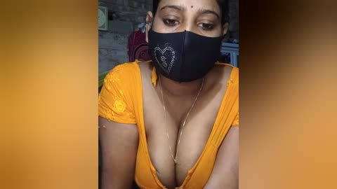 Video of a South Asian woman with medium brown skin, wearing a black face mask with heart design, a low-cut orange top revealing cleavage, and a gold necklace, in a brick-walled room.