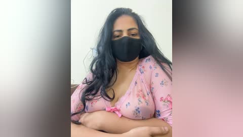 Video of a South Asian woman with long black hair, wearing a pink floral top, black mask, and a pink bow, sitting with crossed arms, against a blurred background.