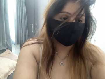 A video of a woman with long, straight, brown hair, wearing a black mask, beige tank top, and a silver necklace, standing indoors near a bed with gray curtains.