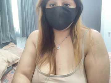 Video of a middle-aged woman with long, straight brown hair, wearing a black face mask, beige spaghetti strap tank top, and a silver necklace, sitting in a dimly lit bedroom with grey curtains and a bed.