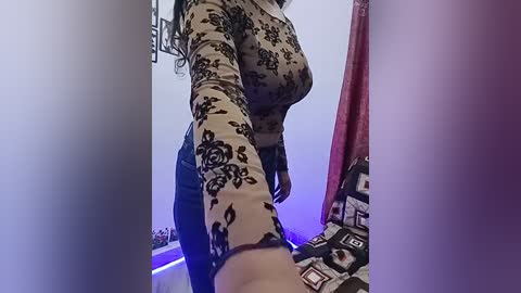 Video of a woman with long, wavy black hair, wearing a tight, black floral-patterned top that accentuates her large breasts. She stands in a dimly lit room with a bed adorned with patterned sheets and a purple curtain.