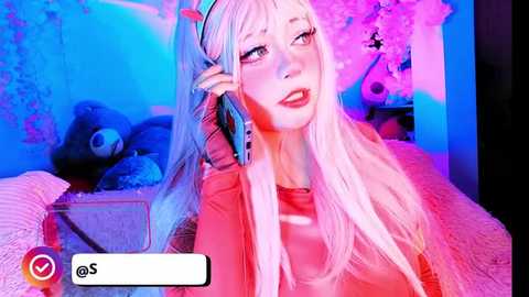Video of a fair-skinned woman with long, pink hair, red lipstick, and a headset, sitting on a bed with plush toys and a cherry blossom backdrop, bathed in blue and pink lighting.