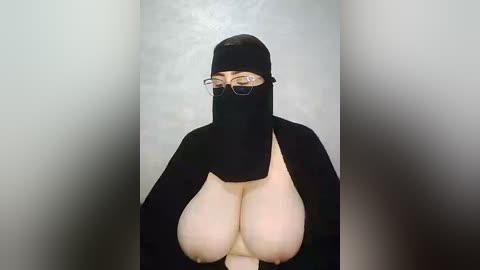Video of a pale-skinned woman in a black niqab and glasses, revealing a large, bare, pendulous breast. She stands against a plain, light gray background, emphasizing her head covering and exposed breast.