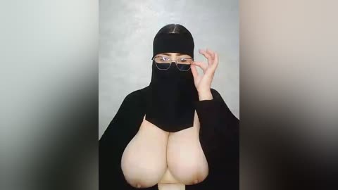 Video of a light-skinned woman with large breasts, wearing a black burqa and glasses, adjusting her glasses. She stands against a plain, light gray wall.