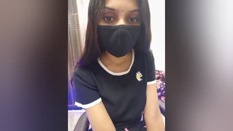 Video of a young South Asian woman with long, straight dark hair, wearing a black face mask, black T-shirt with a sunflower, and sitting indoors.