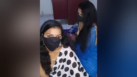 Video of two South Asian women, one with a black mask, the other with a blue mask, sitting in a kitchen with red cabinets and white walls, having their hair braided.