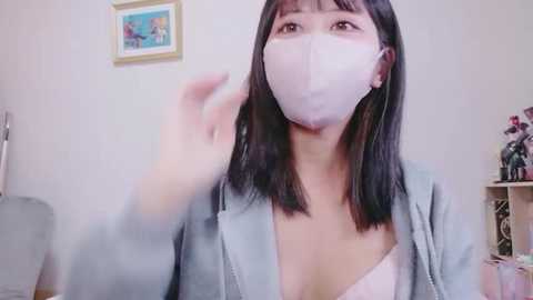 Video of an Asian woman with shoulder-length black hair, wearing a light blue jacket over a white bra, a pink face mask, and a white surgical mask. She is indoors with a framed painting on the wall and a toy shelf in the background.