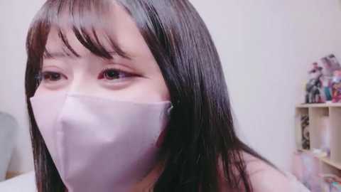 A video of a young Asian woman with long black hair and pale skin, wearing a white surgical mask, sitting indoors with a shelf of colorful figurines in the background.