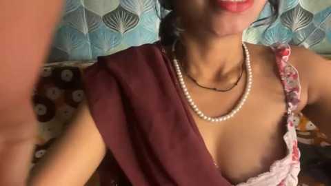 Video of a woman with medium skin tone, dark hair, wearing a maroon dress, pearl necklace, and pearl earrings, smiling. Background features a patterned couch with a blue and white geometric design.