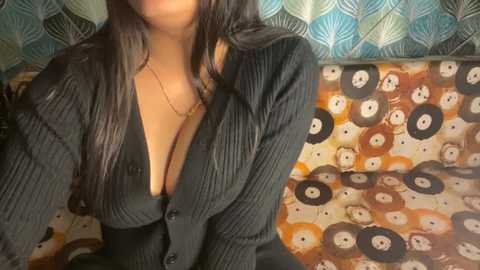 Video of a woman with medium brown skin, long dark hair, wearing a black ribbed cardigan, revealing cleavage, against a backdrop with a patterned cushion and blue, leaf-patterned wallpaper.