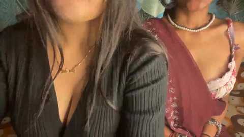 A video shows two women with medium brown skin, wearing a black V-neck sweater and a maroon blouse with lace, respectively. Both have long hair, with the woman in the background wearing a pearl necklace.
