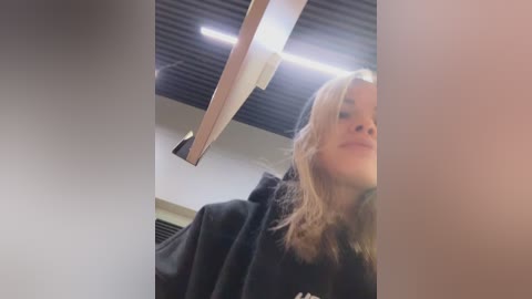 Video of a blonde woman with shoulder-length hair, wearing a black jacket, taken from a low angle. The background features a modern, dark wooden ceiling with a fluorescent light fixture.