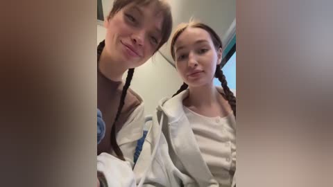 A video of two young girls with light skin and long brown hair in pigtails, smiling while seated in a car, wearing casual clothes and car seat belts.