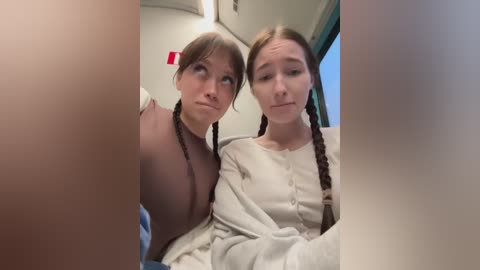 Video of two young women with long brown hair in braids, wearing white sweaters, seated on a bus. One woman has a subtle, almost pensive expression, while the other looks neutral.