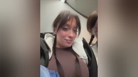 Video of a woman with light skin, brown hair, and bangs, wearing a black jacket and brown top, smiling. Background shows airplane seats and passengers.