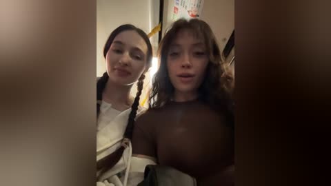 A video shows two young women with long hair, one with braids, standing close in a dimly lit room. One woman is topless, revealing large breasts.