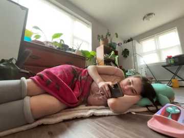 Video of a young woman in a pink towel, lying on a wooden floor with plants and a computer nearby in a bright, cluttered room.