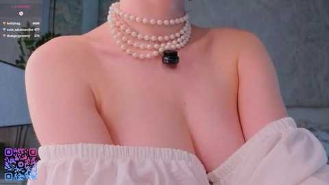 A video of a light-skinned woman with a large bust wearing an off-shoulder white top, pearl necklace, and black pendant. The background is a blurred, modern room with a green wall.